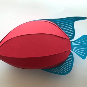 May include: A three-dimensional paper craft fish featuring a rounded, segmented red body. The fish has bright blue fins and a blue tail, each detailed with thin, parallel cut-out slits that create a striped, open-work appearance.