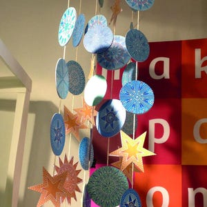 Op de afbeelding: Een kleurrijk mobiel met blauwe, groene en oranje papieren cirkels en sterren die aan een witte ronde basis hangen. Het mobiel hangt aan het plafond en is versierd met ingewikkelde patronen. De tekst "www.pappet.com" is zichtbaar op een rood en roze oppervlak onder het mobiel.