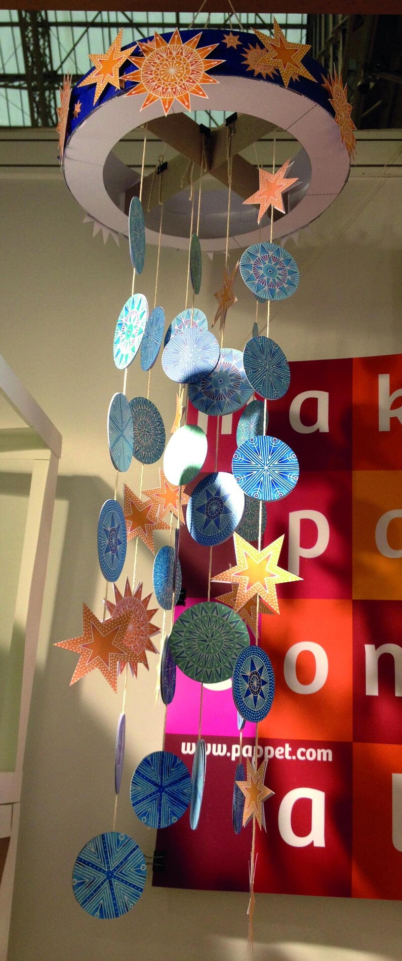 Op de afbeelding: Een kleurrijk mobiel met blauwe, groene en oranje papieren cirkels en sterren die aan een witte ronde basis hangen. Het mobiel hangt aan het plafond en is versierd met ingewikkelde patronen. De tekst "www.pappet.com" is zichtbaar op een rood en roze oppervlak onder het mobiel.