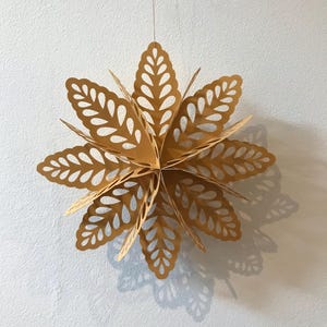 Op de afbeelding: Een driedimensionaal hangend sterornament gemaakt van goudkleurig papier. De structuur bestaat uit meerdere lagen puntige, bladvormige bloemblaadjes met ingewikkelde uitgesneden patronen, wat een symmetrische, stervormige geometrische vorm cre&euml;ert.