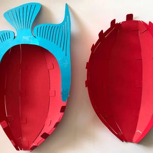 May include: Two red, bowl-shaped containers with a curved, segmented design. One container features a bright blue, decorative fish-shaped attachment on its upper edge, while the other is a plain red, oval-shaped bowl with small tabs along the rim.