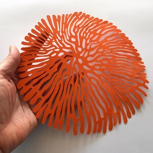May include: A circular orange placemat featuring a complex, organic cut-out pattern that radiates from the center outward. The design consists of numerous thin, branching lines that create a coral-like or brain-like aesthetic across the surface.