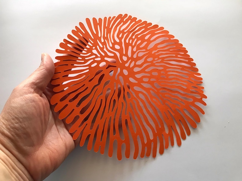May include: A circular orange placemat featuring a complex, organic cut-out pattern that radiates from the center outward. The design consists of numerous thin, branching lines that create a coral-like or brain-like aesthetic across the surface.