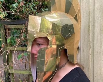 Puede incluir: Un casco de cartón de color dorado con una cresta puntiaguda. El casco está hecho de muchas piezas pequeñas de cartón superpuestas, y tiene una superficie texturizada. El casco está siendo usado por una persona, pero la cara de la persona no es visible.