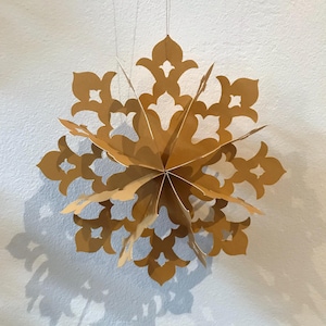 Op de afbeelding: Een driedimensionaal gouden papieren ornament in de vorm van een meerpuntige ster of sneeuwvlok. De structuur bestaat uit verschillende gelaagde, uitgesneden panelen met ingewikkelde, symmetrische bloemachtige patronen, opgehangen aan dunne draden.