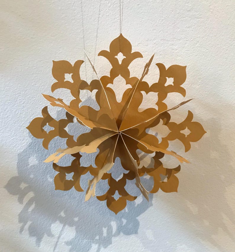 Op de afbeelding: Een driedimensionaal gouden papieren ornament in de vorm van een meerpuntige ster of sneeuwvlok. De structuur bestaat uit verschillende gelaagde, uitgesneden panelen met ingewikkelde, symmetrische bloemachtige patronen, opgehangen aan dunne draden.