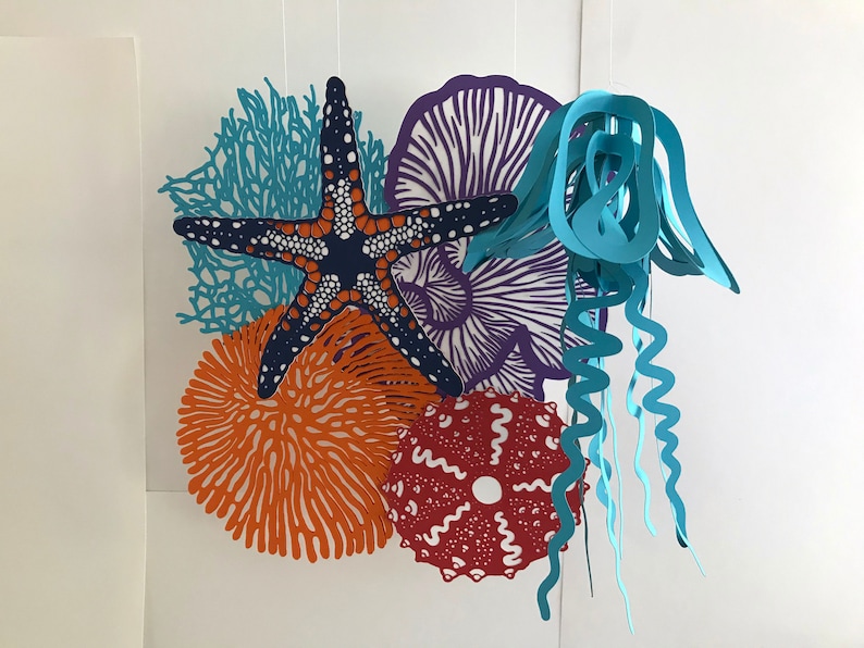 May include: A hanging mobile featuring layered paper cutouts of marine life, including a dark blue starfish with white spots, a light blue jellyfish, a bright orange sea anemone, a purple coral fan, and a textured red sea urchin shape.