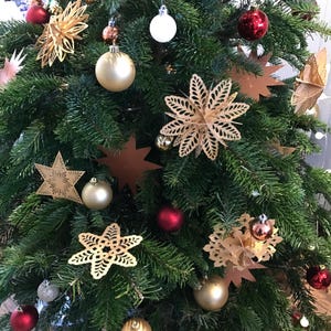 Op de afbeelding: Een groene kerstboom versierd met diverse ornamenten, waaronder gouden en rode kerstballen, ingewikkelde lasergesneden gouden stervormige decoraties en platte koperkleurige sterren tussen de dichte dennennaalden.