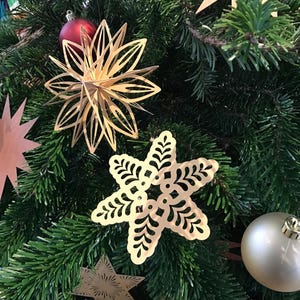 Op de afbeelding: Een verzameling goudkleurige, lasergesneden metalen ornamenten die in een groene dennenboom hangen. De ornamenten bestaan uit een meerlaagse ster, een plat sneeuwvlokvormig stuk met uitsparingen en een ster met stralende lijnen.