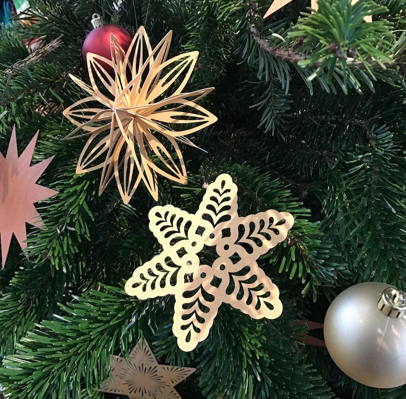 Op de afbeelding: Een verzameling goudkleurige, lasergesneden metalen ornamenten die in een groene dennenboom hangen. De ornamenten bestaan uit een meerlaagse ster, een plat sneeuwvlokvormig stuk met uitsparingen en een ster met stralende lijnen.