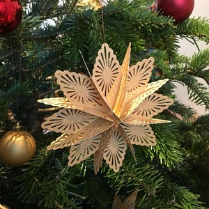 Op de afbeelding: Een driedimensionaal stervormig ornament gemaakt van lichtbeige papier met ingewikkelde lasergesneden patronen. De ster heeft meerdere gelaagde punten die vanuit een centrale as uitstralen, wat een geometrisch ontwerp cre&euml;ert dat aan een dun touwtje hangt.