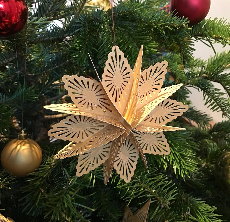 Op de afbeelding: Een driedimensionaal stervormig ornament gemaakt van lichtbeige papier met ingewikkelde lasergesneden patronen. De ster heeft meerdere gelaagde punten die vanuit een centrale as uitstralen, wat een geometrisch ontwerp cre&euml;ert dat aan een dun touwtje hangt.