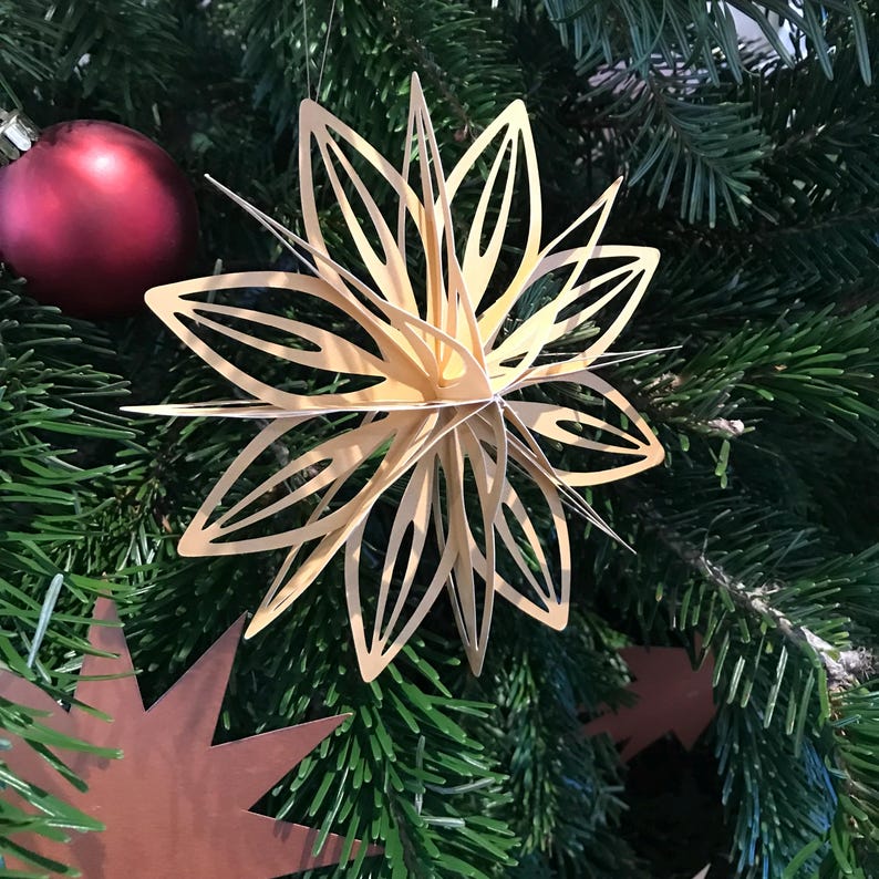 Op de afbeelding: Een driedimensionaal stervormig ornament gemaakt van lichtbeige papier met meerdere puntige bloemblaadjes die vanuit een centraal punt uitstralen, hangend tussen groene dennennaalden naast een matte rode kerstbal.