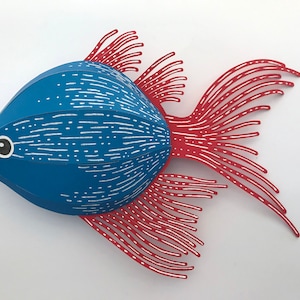May include: A paper craft fish with a rounded blue body featuring white horizontal dashed lines and a black and white eye. The fish has long, thin, red fins and a tail with white linear details, creating a layered and textured appearance.