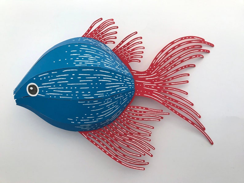 May include: A paper craft fish with a rounded blue body featuring white horizontal dashed lines and a black and white eye. The fish has long, thin, red fins and a tail with white linear details, creating a layered and textured appearance.