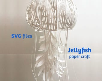 May include: A white paper craft jellyfish with a dome-shaped top featuring intricate cut-out patterns and long, flowing paper tentacles hanging downward. The text "SVG files" and "Jellyfish paper craft" is printed in blue on the image.
