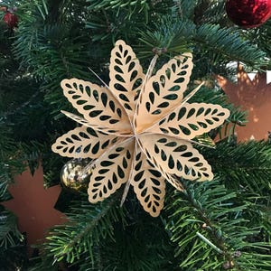 Op de afbeelding: Een driedimensionaal stervormig ornament gemaakt van lichtgoud papier met ingewikkelde bladvormige uitsparingen. Het meerlaagse ontwerp heeft puntige bloemblaadjes die vanuit een centraal punt uitstralen, opgehangen aan een dunne draad.