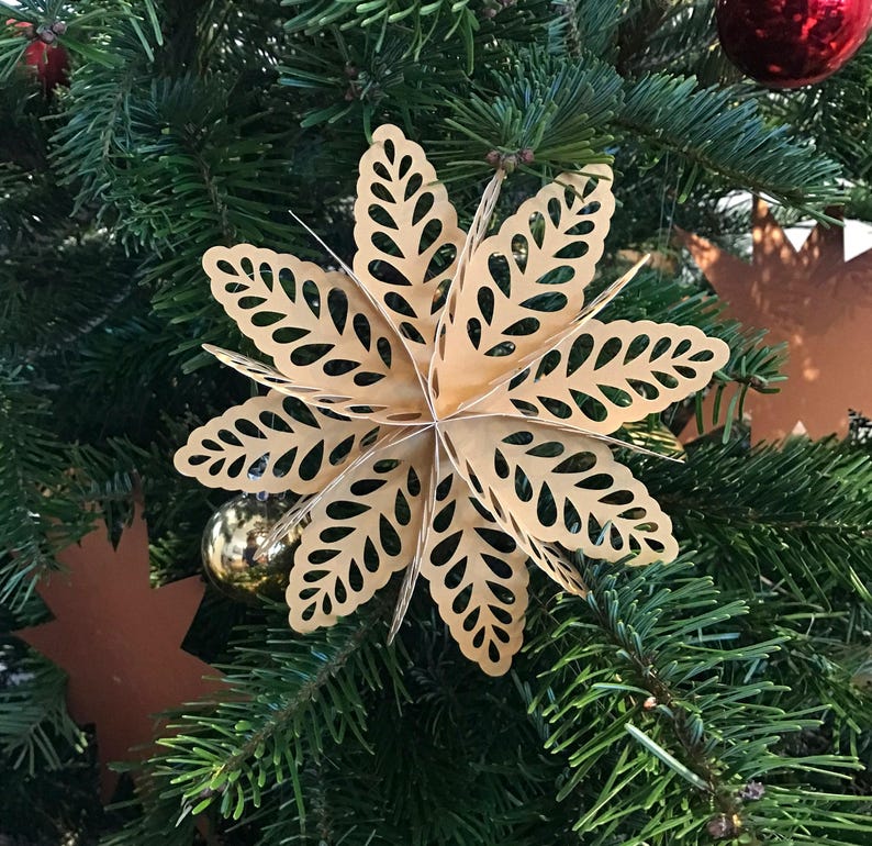 Op de afbeelding: Een driedimensionaal stervormig ornament gemaakt van lichtgoud papier met ingewikkelde bladvormige uitsparingen. Het meerlaagse ontwerp heeft puntige bloemblaadjes die vanuit een centraal punt uitstralen, opgehangen aan een dunne draad.