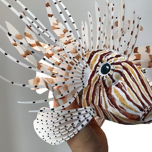 May include: A paper model of a lionfish with white, brown, and black stripes and spots. The lionfish has a large, fan-like fin on its back and a smaller fin on its belly.