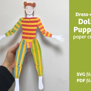 May include: A paper doll puppet with a striped top in red, orange, and yellow, and striped green and blue legs. The text on the right says "Dress-up Doll Puppet paper craft". The doll's limbs are articulated. A hand holds the doll.