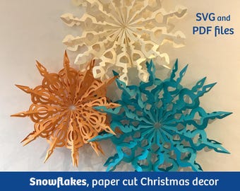 May include: Three paper snowflakes in orange, white, and blue. The snowflakes are layered on top of each other. The text "SVG and PDF files" is in the top right corner of the image. The text "Snowflakes, paper cut Christmas decor" is at the bottom of the image.