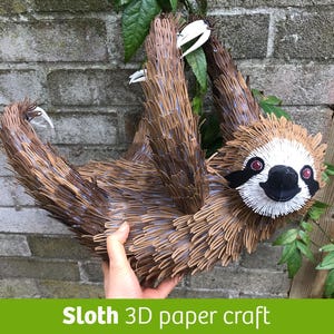 Puede incluir: Un perezoso artesanal de papel 3D en tonos marrones y beige, colgado de una rama. El perezoso tiene una cara blanca, detalles negros y ojos rojos. El texto "Sloth 3D paper craft" está en un rectángulo verde.