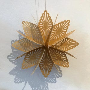Op de afbeelding: Een goudkleurig, meerpuntig sterornament gemaakt van gevouwen papier. De ster is voorzien van ingewikkelde, symmetrische uitgesneden patronen op elk van de punten, wat zorgt voor een delicaat, kantachtig uiterlijk. Het hangt aan een dun, lichtgekleurd touwtje.