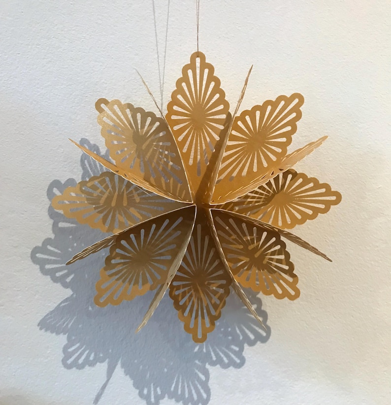 Op de afbeelding: Een goudkleurig, meerpuntig sterornament gemaakt van gevouwen papier. De ster is voorzien van ingewikkelde, symmetrische uitgesneden patronen op elk van de punten, wat zorgt voor een delicaat, kantachtig uiterlijk. Het hangt aan een dun, lichtgekleurd touwtje.