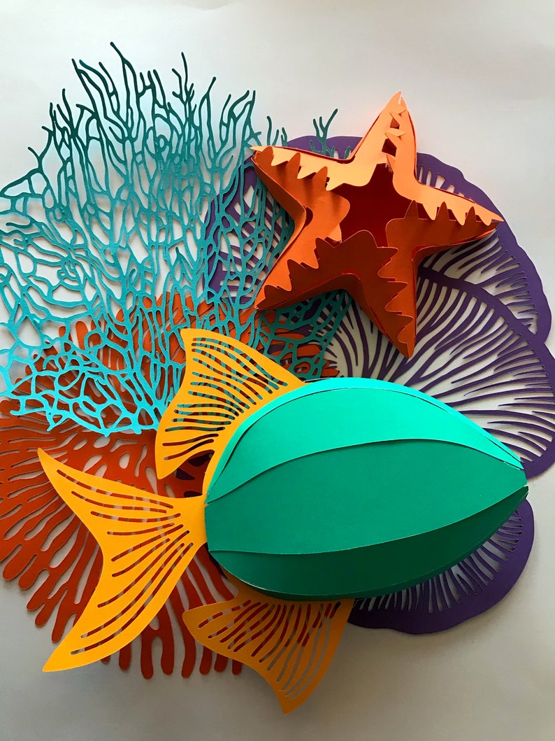 May include: A collection of paper cutouts featuring a three-dimensional orange starfish, a three-dimensional green fish with orange fins, and flat, intricate coral-like shapes in teal, orange, and purple, all arranged in a layered composition.
