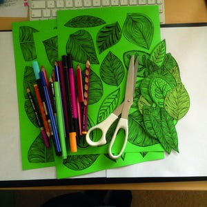 May include: Three green sheets of paper with black line drawings of leaves. A pair of silver scissors and a variety of colored markers are on top of the sheets.