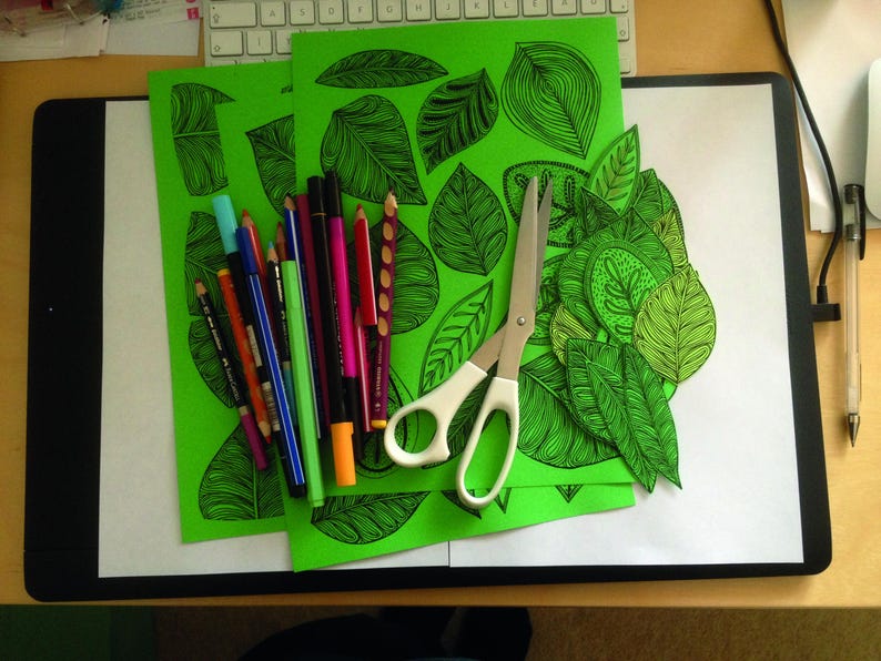 May include: Three green sheets of paper with black line drawings of leaves. A pair of silver scissors and a variety of colored markers are on top of the sheets.