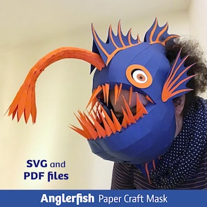 Op de afbeelding: Een blauw en oranje papieren knutselmasker in de vorm van een zeeduivel met scherpe tanden, een lange lokhengel en vinnen. Het masker heeft een opvallend oog en gestructureerde segmenten. De tekst luidt: SVG and PDF files, Anglerfish Paper Craft Mask.