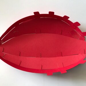 May include: A bright red paper craft bowl with an oval, football-like shape. The structure is composed of multiple interlocking paper segments with small tabs along the edges, creating a layered, open-top container with a smooth interior.