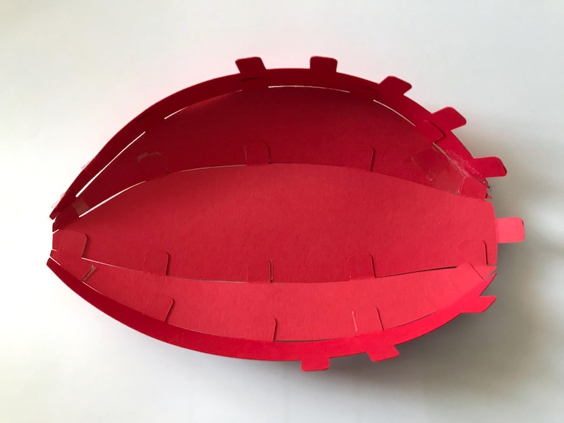 May include: A bright red paper craft bowl with an oval, football-like shape. The structure is composed of multiple interlocking paper segments with small tabs along the edges, creating a layered, open-top container with a smooth interior.