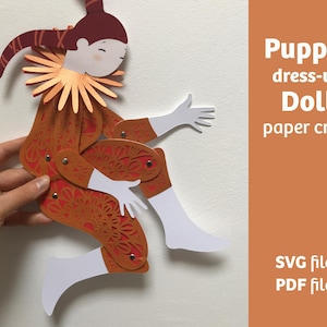 May include: A paper puppet doll in shades of red and orange, with articulated joints and a decorative collar. The image includes the text "Puppet dress-up Doll paper craft" and mentions SVG and PDF files.