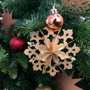Op de afbeelding: Een driedimensionale gouden papieren sneeuwvlokornament met ingewikkelde uitgesneden patronen hangend aan een groene dennentak, vergezeld door een glanzend koperkleurig bolvormig ornament en een mat rood bolvormig ornament.