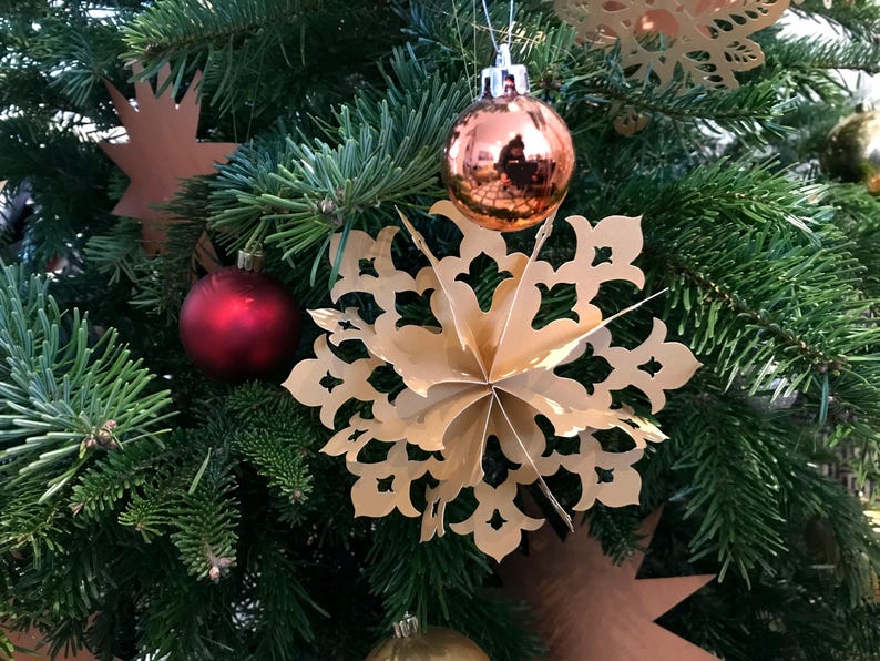 Op de afbeelding: Een driedimensionale gouden papieren sneeuwvlokornament met ingewikkelde uitgesneden patronen hangend aan een groene dennentak, vergezeld door een glanzend koperkleurig bolvormig ornament en een mat rood bolvormig ornament.