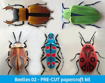 May include: A collection of five papercraft beetles from the Beetles 02 - PRE-CUT papercraft kit. The insects feature vibrant colors including orange, green, yellow, blue, and red with distinct patterns, black legs, and antennae on a white background.