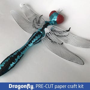 May include: A dragonfly pre-cut paper craft kit featuring a metallic blue and black body with large red eyes and intricate, silver-coloured, laser-cut wings with detailed vein patterns. The text "Dragonfly, PRE-CUT paper craft kit" is visible.