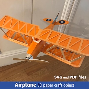 Op de afbeelding: Een oranje dubbeldekker 3D-papieren knutselobject met een zilveren propeller en blauw-witte cirkelvormige markeringen op de staartvleugels. De tekst SVG and PDF files en Airplane 3D paper craft object is onderaan zichtbaar.