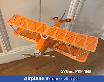 May include: An orange biplane 3D paper craft object with a silver propeller and blue and white circular markings on the tail wings. The text SVG and PDF files and Airplane 3D paper craft object are visible at the bottom of the image.