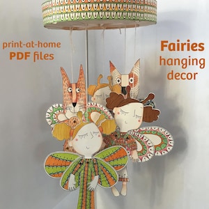 Può includere: Una giostrina sospesa con figure di carta illustrate di fate e una volpe appese a una cornice circolare. Il testo "print-at-home PDF files" e "Fairies hanging decor" è visibile. Le figure presentano motivi nei colori arancione, verde e marrone.