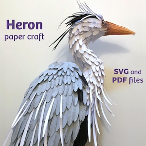 May include: A paper craft heron sculpture featuring layered gray and white feathers, a long neck, and a pointed orange beak. The text Heron paper craft and SVG and PDF files is visible on the sides of the bird against a plain white background.