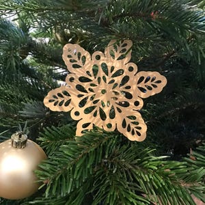 Op de afbeelding: Een platte, goudkleurige sneeuwvlokornament met ingewikkelde, symmetrische uitgesneden patronen. De sneeuwvlok heeft zes hoofdpunten met bladachtige details en cirkelvormige accenten. Een gladde, ronde, goudkleurige bal is ernaast zichtbaar.