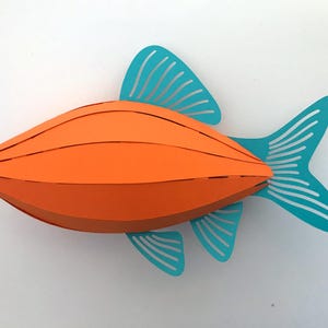 May include: A three-dimensional paper craft fish featuring a bright orange segmented body with a curved, layered design. The fish has teal-colored fins and a tail, each detailed with thin, parallel cut-out slits that create a striped appearance.