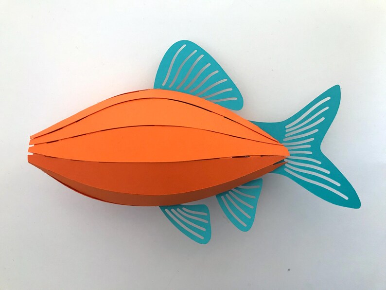 May include: A three-dimensional paper craft fish featuring a bright orange segmented body with a curved, layered design. The fish has teal-colored fins and a tail, each detailed with thin, parallel cut-out slits that create a striped appearance.