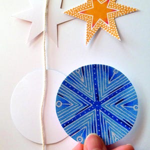 Op de afbeelding: Een string met papieren ornamenten met een witte ster, een gele ster met witte stippen en een blauwe en witte cirkel met een geometrisch patroon.