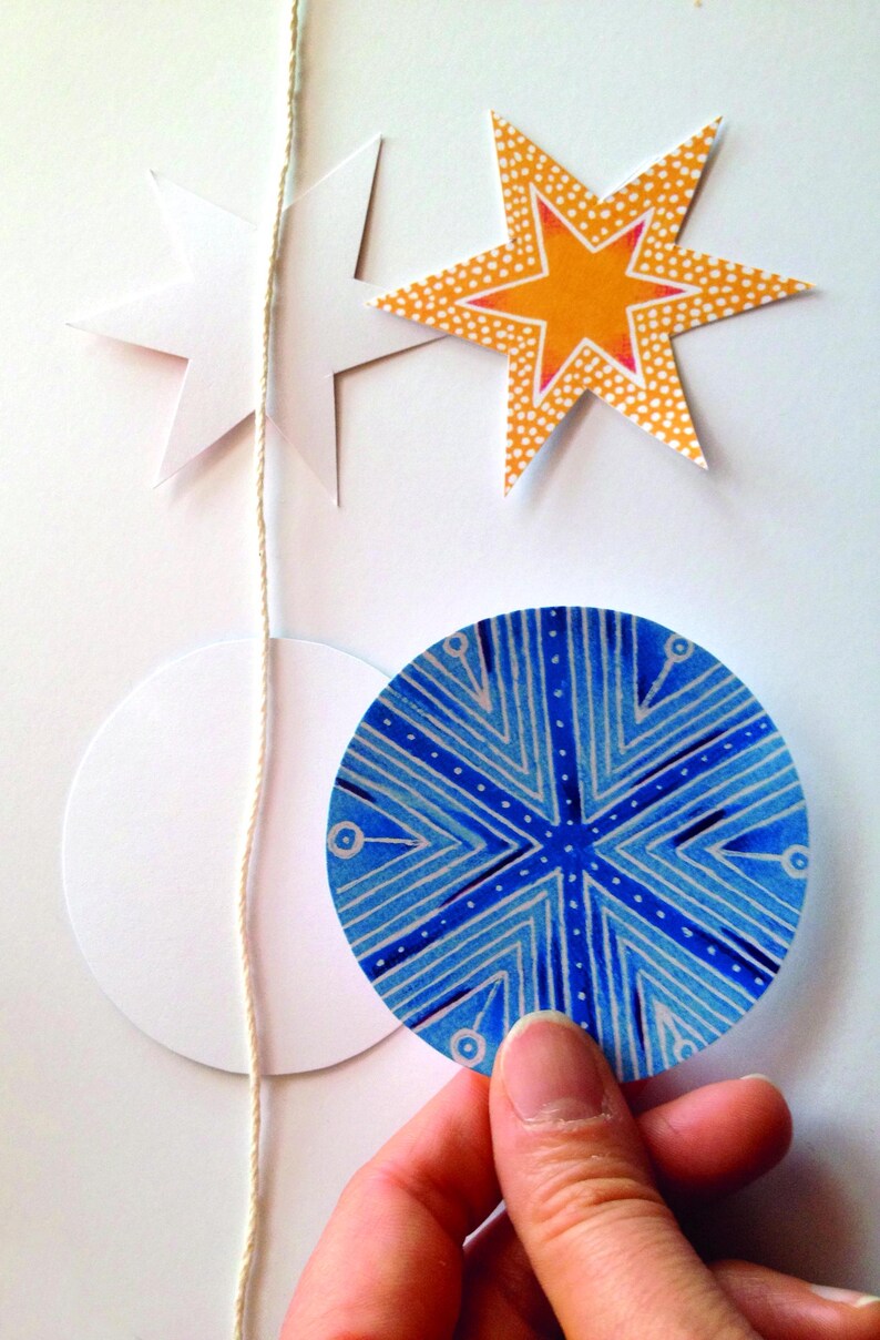 Op de afbeelding: Een string met papieren ornamenten met een witte ster, een gele ster met witte stippen en een blauwe en witte cirkel met een geometrisch patroon.