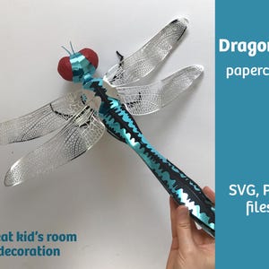Könnte beinhalten: Eine Papierdrachenfliege mit silbernen Flügeln und einem türkisfarbenen und schwarzen Körper. Die Libelle hat einen roten Kopf und ist mit "Dragonfly papercraft" und "Great kid's room decoration" beschriftet. Das Bild enthält auch den Text "SVG, PDF files".