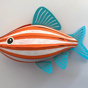 May include: A three-dimensional paper craft fish sculpture featuring alternating horizontal stripes of orange and white. The fish has a rounded body shape with light blue die-cut fins and a tail, and a single black and white circular eye detail.